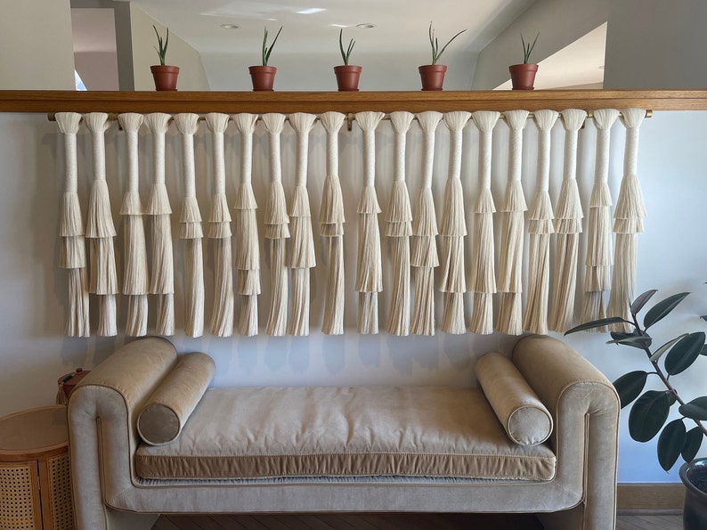 K&ouml;nnte beinhalten: Ein wei&szlig;er Makramee-Wandbehang mit mehreren Reihen von Quasten, der &uuml;ber einem hellbraunen Sofa mit zwei Kissen h&auml;ngt. Der Wandbehang ist an einem Holzregal mit kleinen Topfpflanzen befestigt.