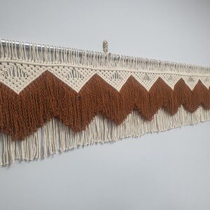 May include: A macrame wall hanging with a geometric design. The piece features a cream-colored top section with a zig-zag pattern in brown, and a fringe of cream-colored rope. The wall hanging is mounted on a metal rod.