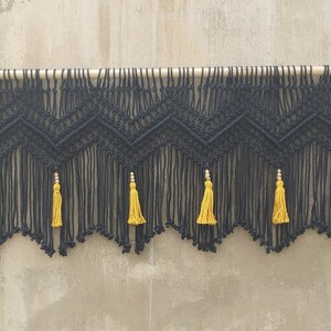 May include: Black macrame wall hanging with a geometric pattern and yellow tassels. The hanging is attached to a wooden dowel rod.