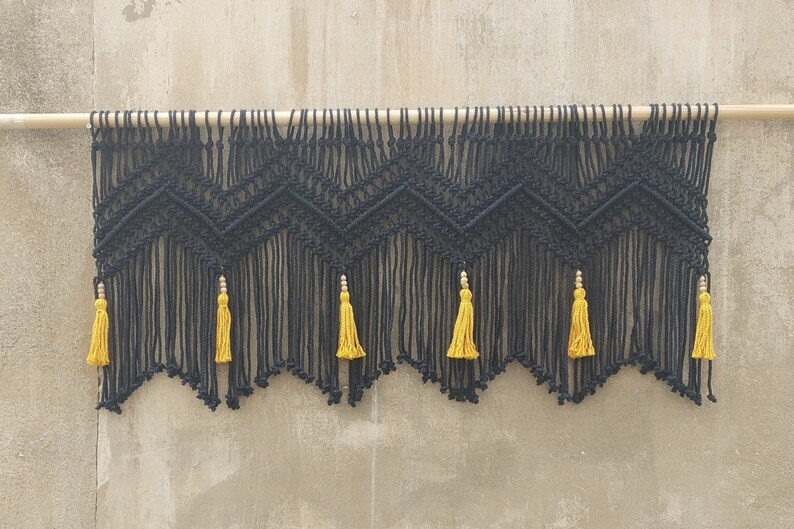 May include: Black macrame wall hanging with a geometric pattern and yellow tassels. The hanging is attached to a wooden dowel rod.