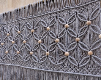 May include: A grey macrame wall hanging featuring a repeating geometric knot pattern suspended from a wooden dowel. The textile design incorporates natural light-colored wooden beads integrated into the center of each knotted diamond shape.