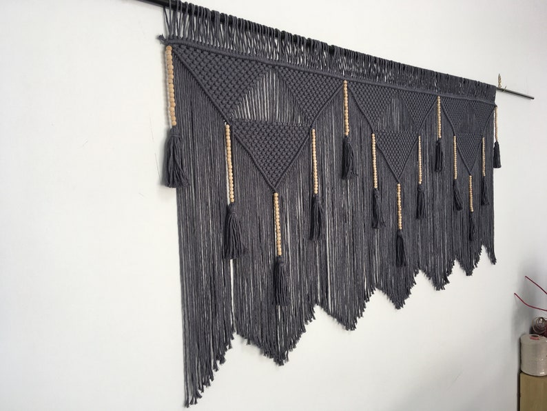 May include: Gray macrame wall hanging with wooden beads and tassels. The hanging features a geometric pattern with a triangular shape.