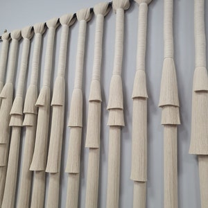 K&ouml;nnte beinhalten: Ein Wandbehang aus Naturfaserseil mit mehreren Fransenreihen. Das Seil ist hellbeige und an einer schwarzen Stange aufgeh&auml;ngt.