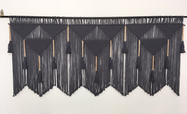 May include: Gray macrame wall hanging with wooden beads and tassels. The wall hanging is made of a thick, textured cord and has a geometric pattern.