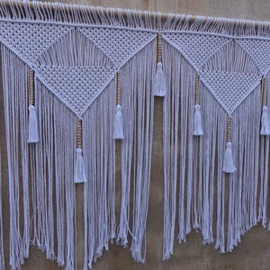 Peut inclure: Une suspension murale en macramé blanche avec un motif géométrique et des perles en bois. La suspension est composée de trois sections avec des glands en bas.