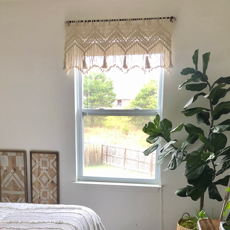 K&ouml;nnte beinhalten: Ein wei&szlig;er Makramee-Fenster&uuml;berhang mit Quasten h&auml;ngt &uuml;ber einem Fenster mit Blick auf einen gr&uuml;nen Garten und B&auml;ume. Eine gro&szlig;e gr&uuml;ne Topfpflanze steht in der Ecke des Raumes.