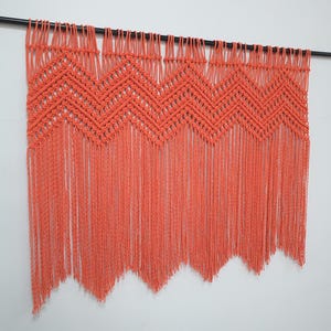 May include: Coral-colored macrame wall hanging. The handmade piece features a geometric chevron design across the top, with long fringe hanging below. The wall decor is suspended from a black rod.