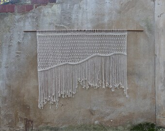May include: A white macrame wall hanging with a wavy pattern and fringe. The hanging is mounted on a wall with a wooden dowel.