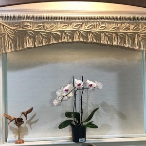 Könnte beinhalten: Ein cremefarbener Makramee-Volant mit Blättermuster hängt über einem Fenster. Darunter befindet sich eine weiße Orchidee mit violetten Zentren in einem schwarzen Topf. Links befindet sich eine Holzskulptur von Vögeln.