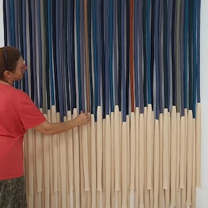 May include: A wall hanging made of yarn in various shades of blue, green, and beige. The yarn is woven together to create a striped pattern. The hanging is approximately 3 feet wide and 4 feet tall.