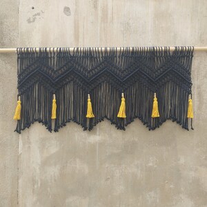 May include: Black macrame wall hanging with a geometric pattern and yellow tassels. The hanging is attached to a wooden dowel rod.