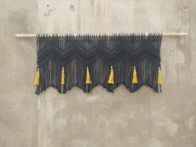 May include: Black macrame wall hanging with a geometric pattern and yellow tassels. The hanging is attached to a wooden dowel rod.