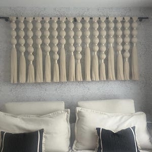 May include: A white macrame wall hanging with a series of knotted balls and tassels. The hanging is attached to a black rod and is displayed on a white wall.