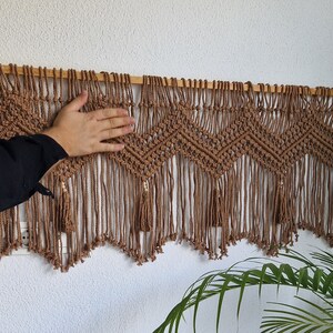 May include: A brown macrame wall hanging with a wood dowel rod at the top. The hanging features a geometric pattern with tassels at the bottom.