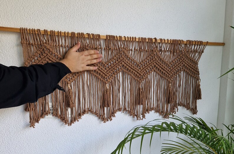 May include: A brown macrame wall hanging with a wood dowel rod at the top. The hanging features a geometric pattern with tassels at the bottom.