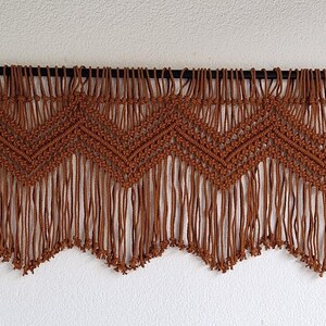 May include: A brown macrame wall hanging with a chevron pattern and fringe. The hanging is made of thick cord and has a rustic, bohemian style.