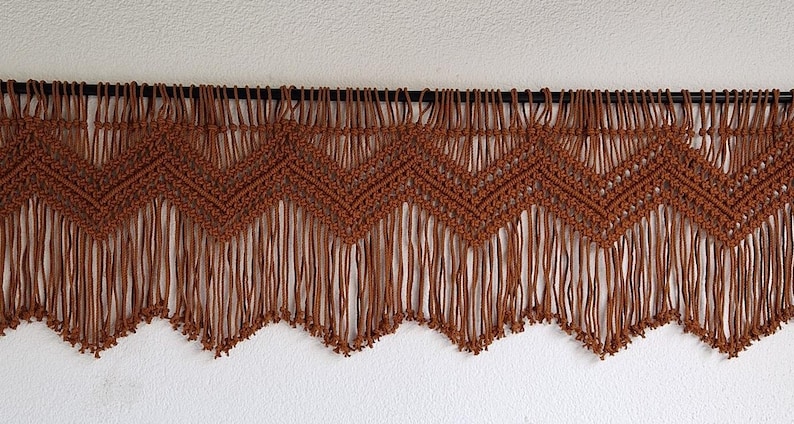 May include: A brown macrame wall hanging with a chevron pattern and fringe. The hanging is made of thick cord and has a rustic, bohemian style.