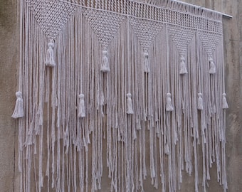 May include: A white macrame wall hanging with a geometric pattern and tassels. The hanging is made of cotton cord and is approximately 3 feet wide and 4 feet long.