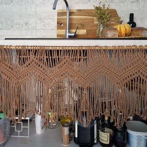 May include: A brown macrame curtain with fringe hangs over a kitchen sink. The curtain is made of natural fibers and has a geometric pattern. The curtain is hanging from a wooden rod.
