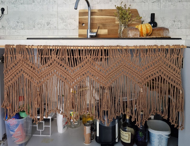 May include: A brown macrame curtain with fringe hangs over a kitchen sink. The curtain is made of natural fibers and has a geometric pattern. The curtain is hanging from a wooden rod.