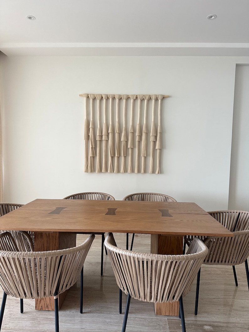 K&ouml;nnte beinhalten: Ein Esstisch aus Holz mit einer nat&uuml;rlichen Holzoptik und einem gewebten Makramee-Wandbehang dar&uuml;ber. Der Tisch hat eine rechteckige Form und ist von sechs gewebten St&uuml;hlen mit schwarzen Metallbeinen umgeben.