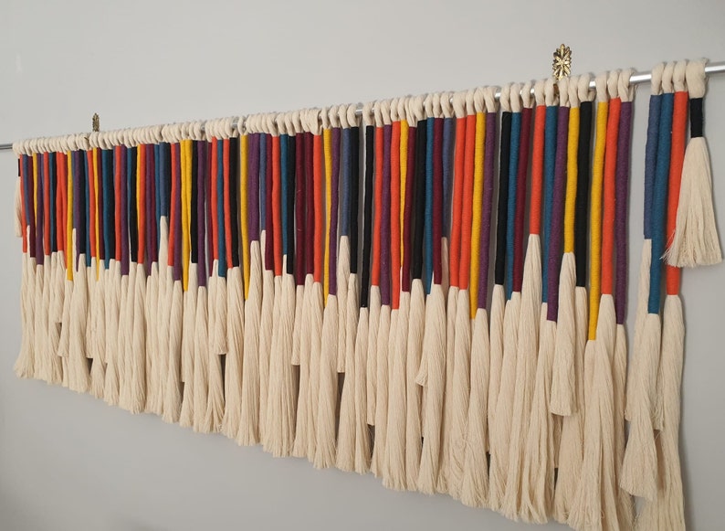 Puede incluir: Un tapiz de macram&eacute; de pared colorido con una variedad de hilos de lana de diferentes colores, incluyendo rojo, naranja, amarillo, verde, azul, morado y negro. Los hilos est&aacute;n atados juntos en la parte superior y cuelgan en cascada de borlas.