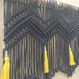 May include: Black macrame wall hanging with a geometric pattern and yellow tassels. The hanging is attached to a wooden dowel.