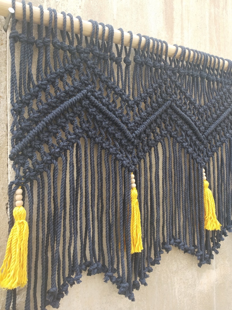 May include: Black macrame wall hanging with a geometric pattern and yellow tassels. The hanging is attached to a wooden dowel.