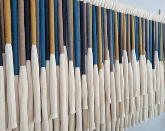 May include: A decorative wall hanging with vertical strands in shades of blue, gold, brown, and grey, topped with white. The strands are arranged in a repeating pattern, creating a textured, decorative piece for the home.