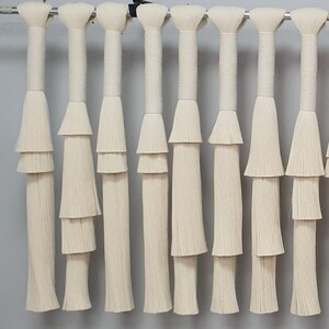 May include: A row of off-white macrame wall hangings. Each hanging features a cylindrical top, a layered, tiered design, and a long, fringed bottom. The neutral color and textured design create a bohemian aesthetic.