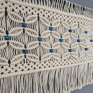 Può includere: Applique da parete in macramè color crema con motivo geometrico e perline blu. Il tessuto fatto a mano presenta un motivo ripetuto di forme a forma di foglia e una frangia nella parte inferiore. Un pezzo decorativo per l'arredamento della casa.