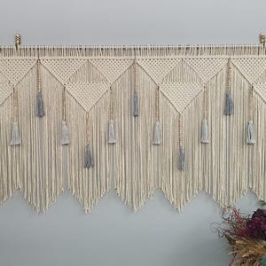 Op de afbeelding: Een macramé wandhanger met een geometrisch patroon en grijze kwastjes. De hanger is gemaakt van natuurlijke vezels en heeft een bohemian stijl.
