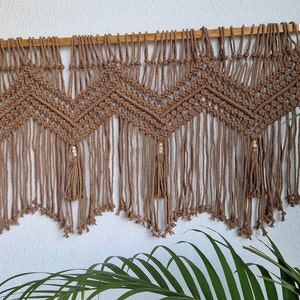 May include: A brown macrame wall hanging with a geometric pattern and wooden beads. The hanging has a fringe at the bottom.