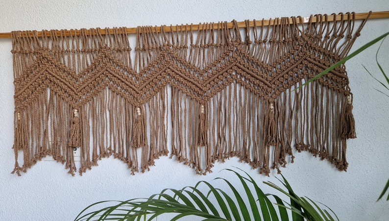 May include: A brown macrame wall hanging with a geometric pattern and wooden beads. The hanging has a fringe at the bottom.