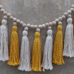 May include: A garland of wooden beads with yellow and white tassels hanging from it. The garland is perfect for adding a touch of bohemian style to your home decor.