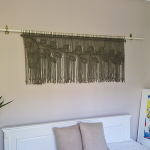 May include: A macrame wall hanging with a leafy design in a muted green color. The hanging is attached to a white rod and is displayed against a light beige wall.