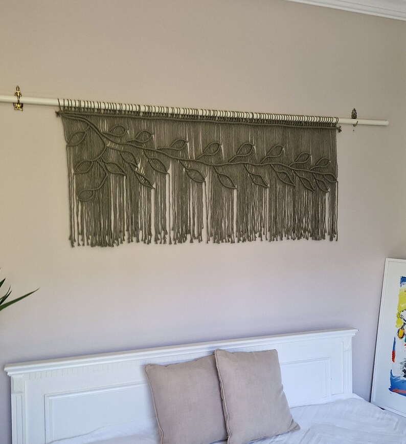 May include: A macrame wall hanging with a leafy design in a muted green color. The hanging is attached to a white rod and is displayed against a light beige wall.