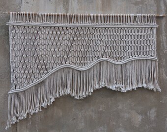 May include: A white macrame wall hanging with a wavy fringe. The hanging is made of natural cotton rope and has a geometric pattern.
