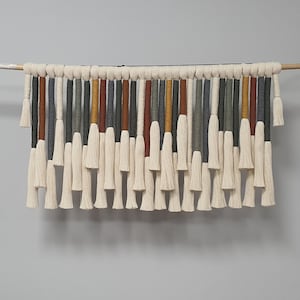 May include: A wall hanging made of white yarn with tassels. The tassels are arranged in a row and are of varying lengths. The yarn is woven with different colored threads, including gray, brown, and yellow.