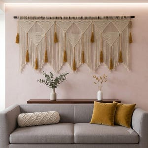 Peut inclure: Une grande tenture murale en macramé blanc cassé suspendue à une tringle horizontale sombre, présentant des motifs géométriques noués complexes, des franges verticales et des pompons jaune moutarde ornés de petites perles en bois.