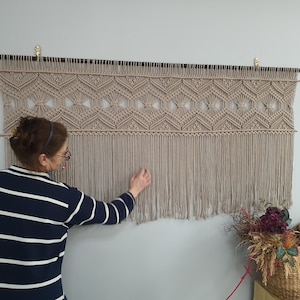 May include: A beige macrame wall hanging with a geometric pattern and fringe. The hanging is mounted on a black rod and is displayed on a white wall.