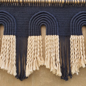 May include: A wall hanging with three woven arches made of dark blue and cream colored rope. The arches are connected by a wooden dowel rod.