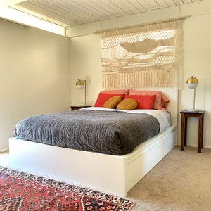 K&ouml;nnte beinhalten: Ein wei&szlig;es Plattformbett mit einer grauen gesteppten Bettdecke und roten und orangefarbenen Kissen. Das Bett steht in einem Schlafzimmer mit einem Makramee-Wandbehang und einem Nachttisch mit einer Lampe.