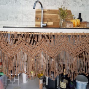 May include: A brown macrame curtain with a geometric pattern hangs from a kitchen cabinet. The curtain has tassels and beads.