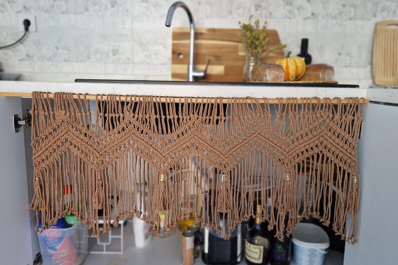 May include: A brown macrame curtain with a geometric pattern hangs from a kitchen cabinet. The curtain has tassels and beads.