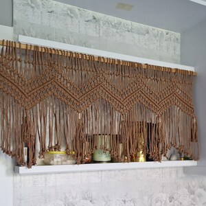 May include: A brown macrame curtain with a geometric pattern hangs above a shelf with various items, including jars and candles.