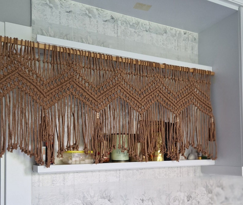 May include: A brown macrame curtain with a geometric pattern hangs above a shelf with various items, including jars and candles.
