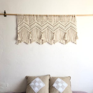 K&ouml;nnte beinhalten: Ein wei&szlig;er Makramee-Wandbehang mit Chevron-Muster und Quasten. Der Behang ist an einer Holzstange befestigt. Zwei beigefarbene Kissen mit wei&szlig;en Spitzenapplikationen liegen auf einem Bett mit einem hellvioletten Bettbezug.