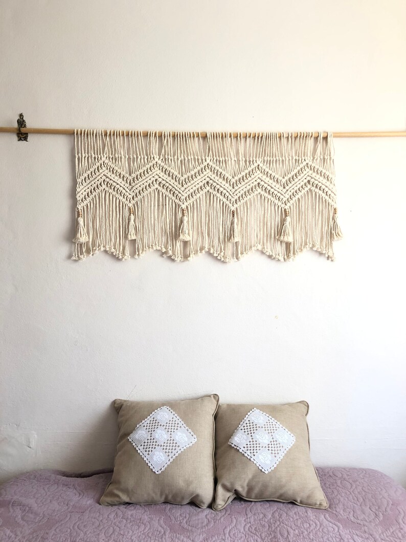 K&ouml;nnte beinhalten: Ein wei&szlig;er Makramee-Wandbehang mit Chevron-Muster und Quasten. Der Behang ist an einer Holzstange befestigt. Zwei beigefarbene Kissen mit wei&szlig;en Spitzenapplikationen liegen auf einem Bett mit einem hellvioletten Bettbezug.