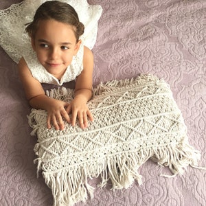 Puede incluir: Una niña pequeña con un vestido blanco de encaje yace sobre una colcha acolchada rosa con una almohada de macramé blanca con flecos.